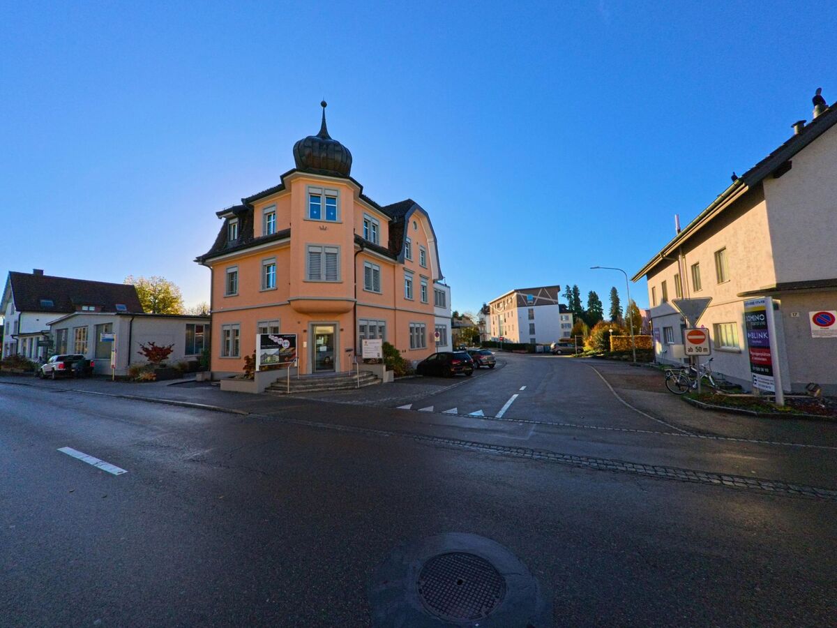 Ein markantes Eckgebaeude mit Turmhaube steht an einer ruhigen Kreuzung in Wil (9500, Kanton St. Gallen) umgeben von Wohnhaeusern bei Sonnenschein.
