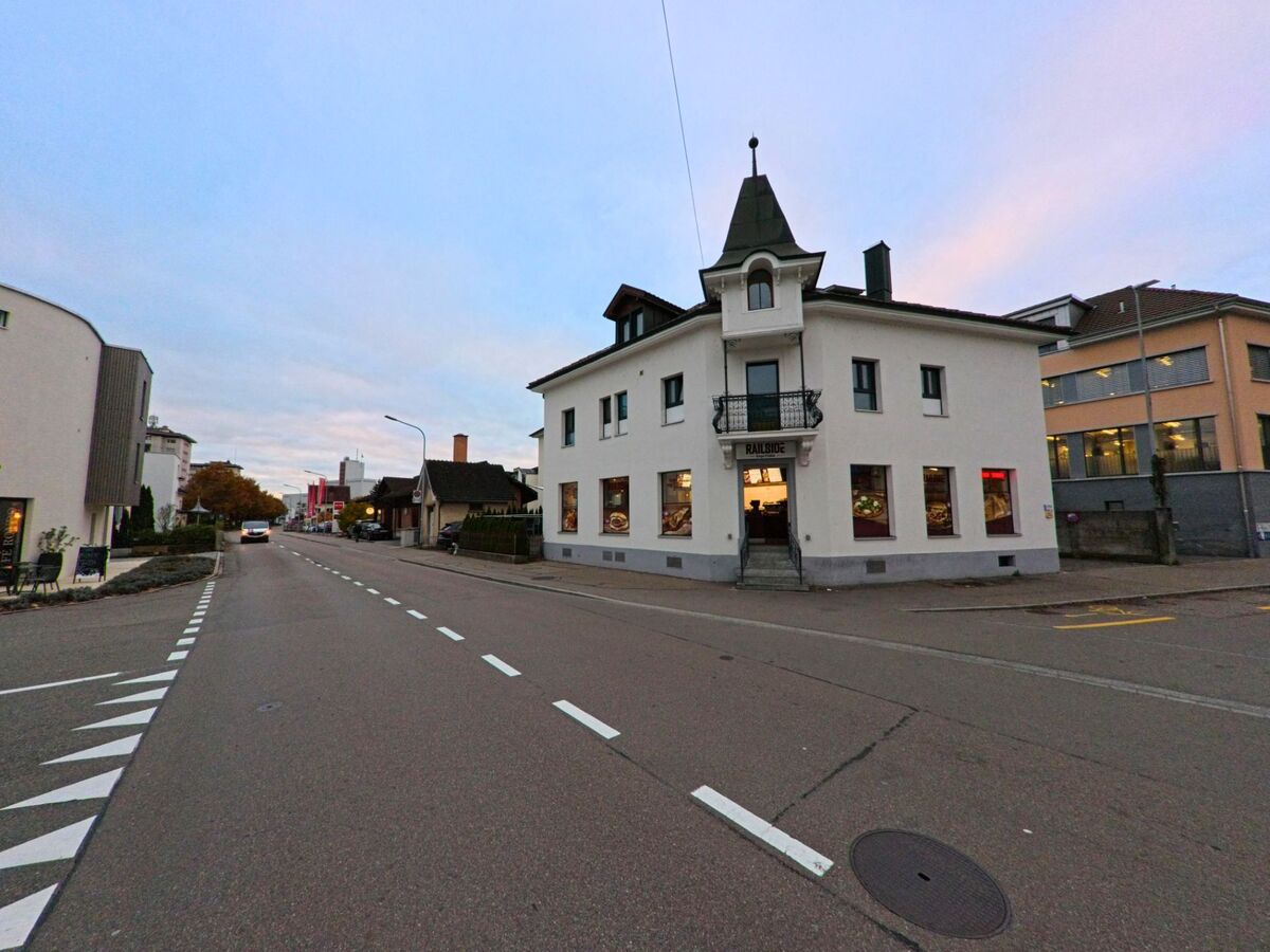 An einer geraden Strasse mit Markierungen steht ein markantes Eckhaus mit Turmaufsatz in Wil (9500, Kanton St. Gallen) zwischen Wohn und Geschaeftsbauten bei Abendlicht.