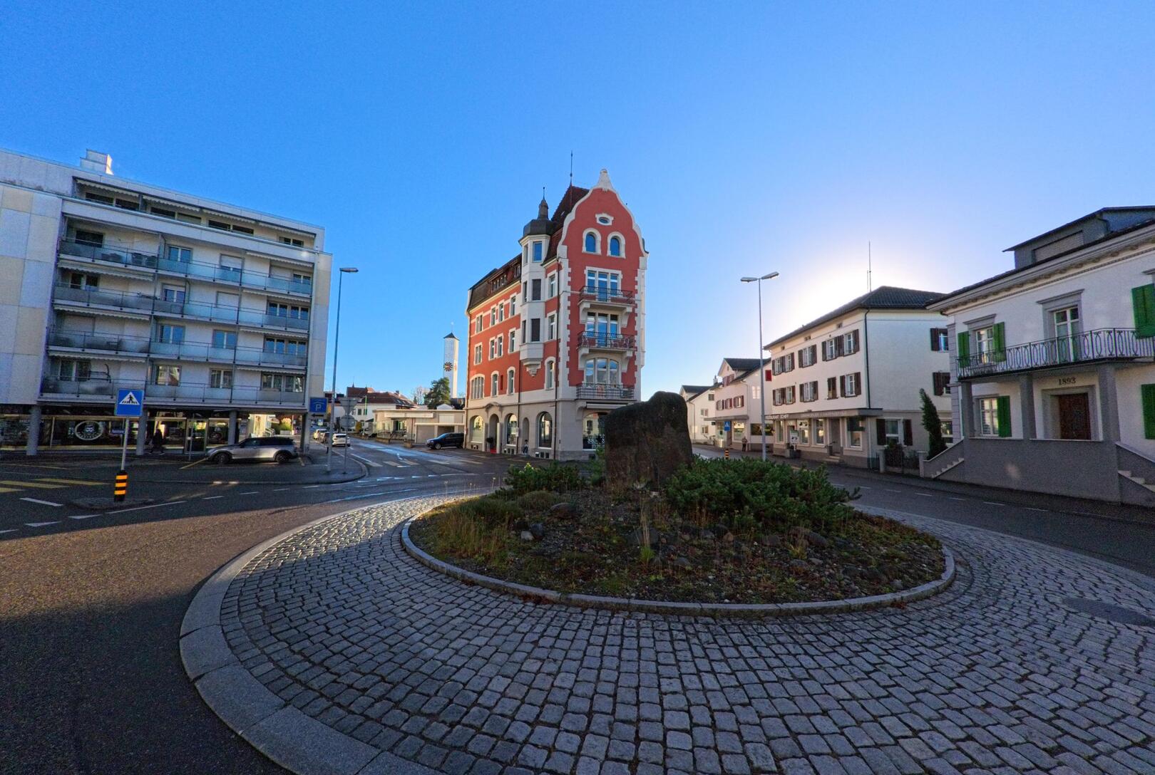 Auf einem gepflasterten Kreisverkehr mit bepflanzter Mittelinsel stehen historische und moderne Gebaeude im Zentrum von Wil (9500, Kanton St. Gallen) bei klarem Himmel.