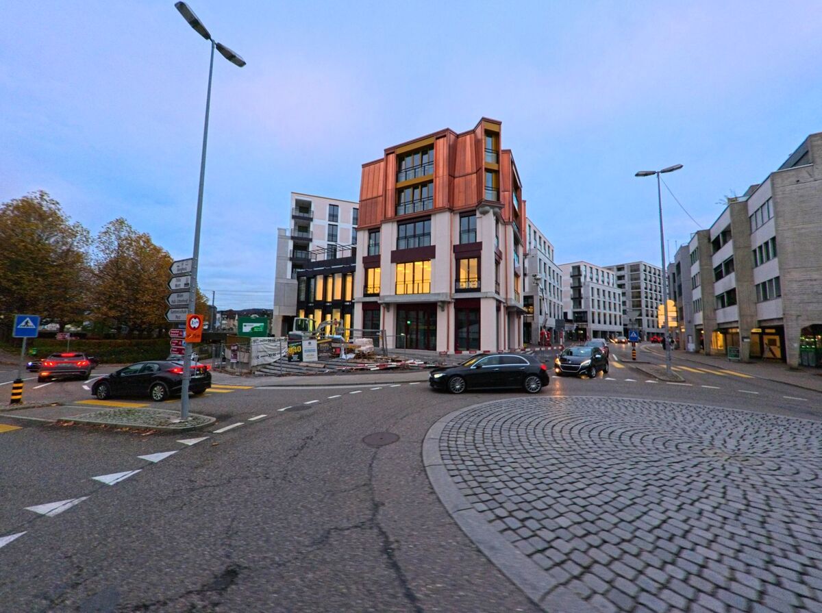 An einer belebten Kreuzung mit Pflasterinsel und Verkehr entsteht ein modernes Gebaeudeensemble in Wil (9500, Kanton St. Gallen) bei einsetzender Abenddaemmerung.
