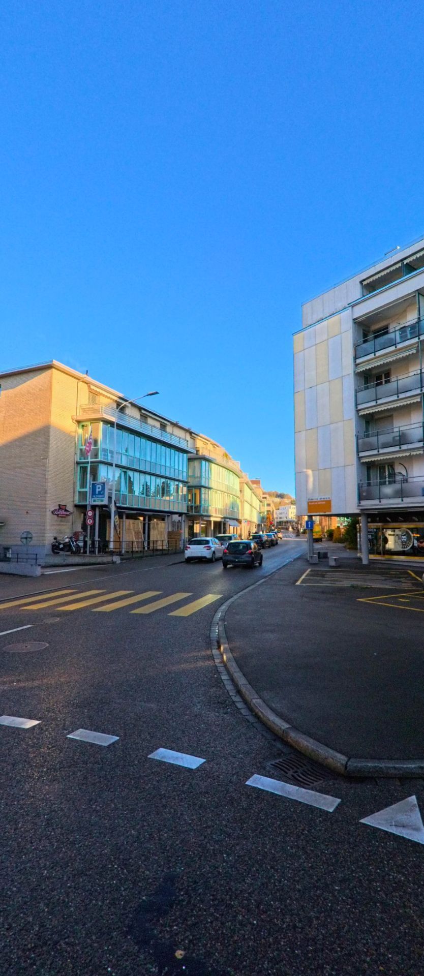 Eine leicht ansteigende Strasse mit Zebrastreifen und modernen Geschaefts und Wohnbauten fuehrt durch ein Stadtquartier von Wil (9500, Kanton St. Gallen) bei klarem Himmel.