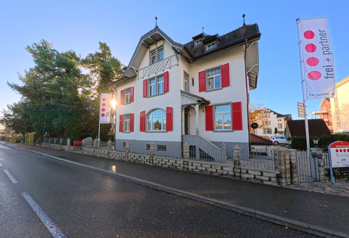 Ein freistehendes Wohnhaus mit roten Fensterlaeden und Vorgarten steht an einer ruhigen Strasse in Wil (9500, Kanton St. Gallen) im warmen Abendlicht.
