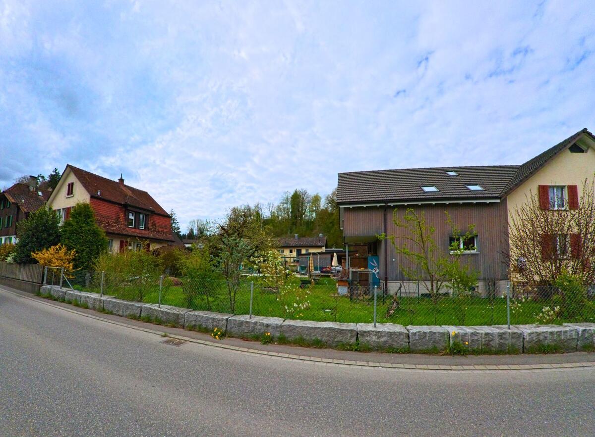 In Wila (8492, Kanton Zürich) führen die Straßen an gepflegten Gärten und Wohnhäusern entlang, mit einer schönen Aussicht auf die Hügel im Hintergrund.