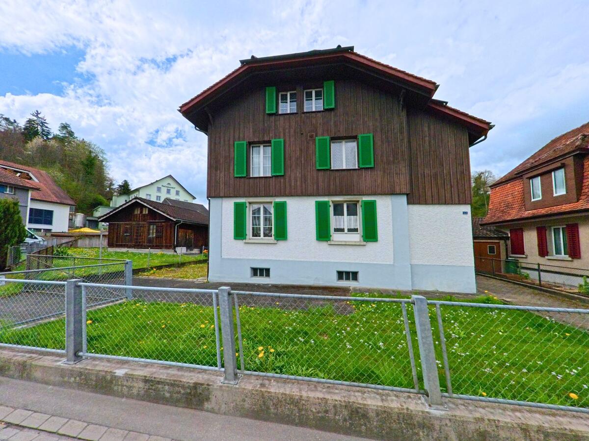 In Wila (8492, Kanton Zürich) ist ein traditionelles Gebäude mit grün gestrichenen Fensterläden zu sehen, das von einem eingezäunten Garten umgeben ist