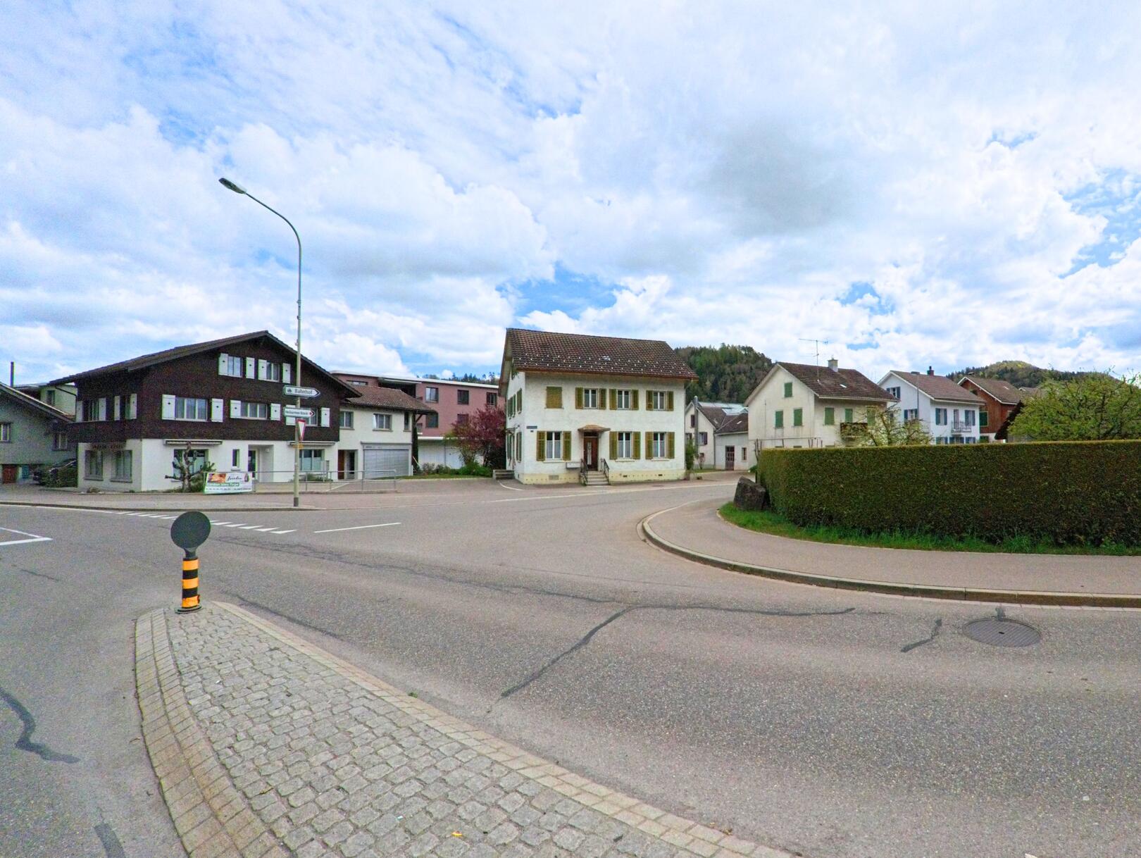 In Wila (8492, Kanton Zürich) trifft eine gepflasterte Verkehrsinsel auf eine Kreuzung mit Wohnhäusern verschiedener Bauweise im Zentrum des Dorfes.