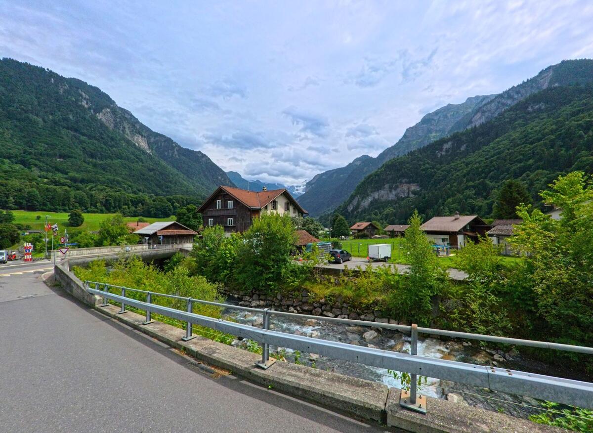 In Wilderswil (3812, Kanton Bern) überquert eine Brücke einen Gebirgsbach, dahinter liegen verstreute Häuser in einem weiten Tal, das von bewaldeten Bergen eingerahmt wird.