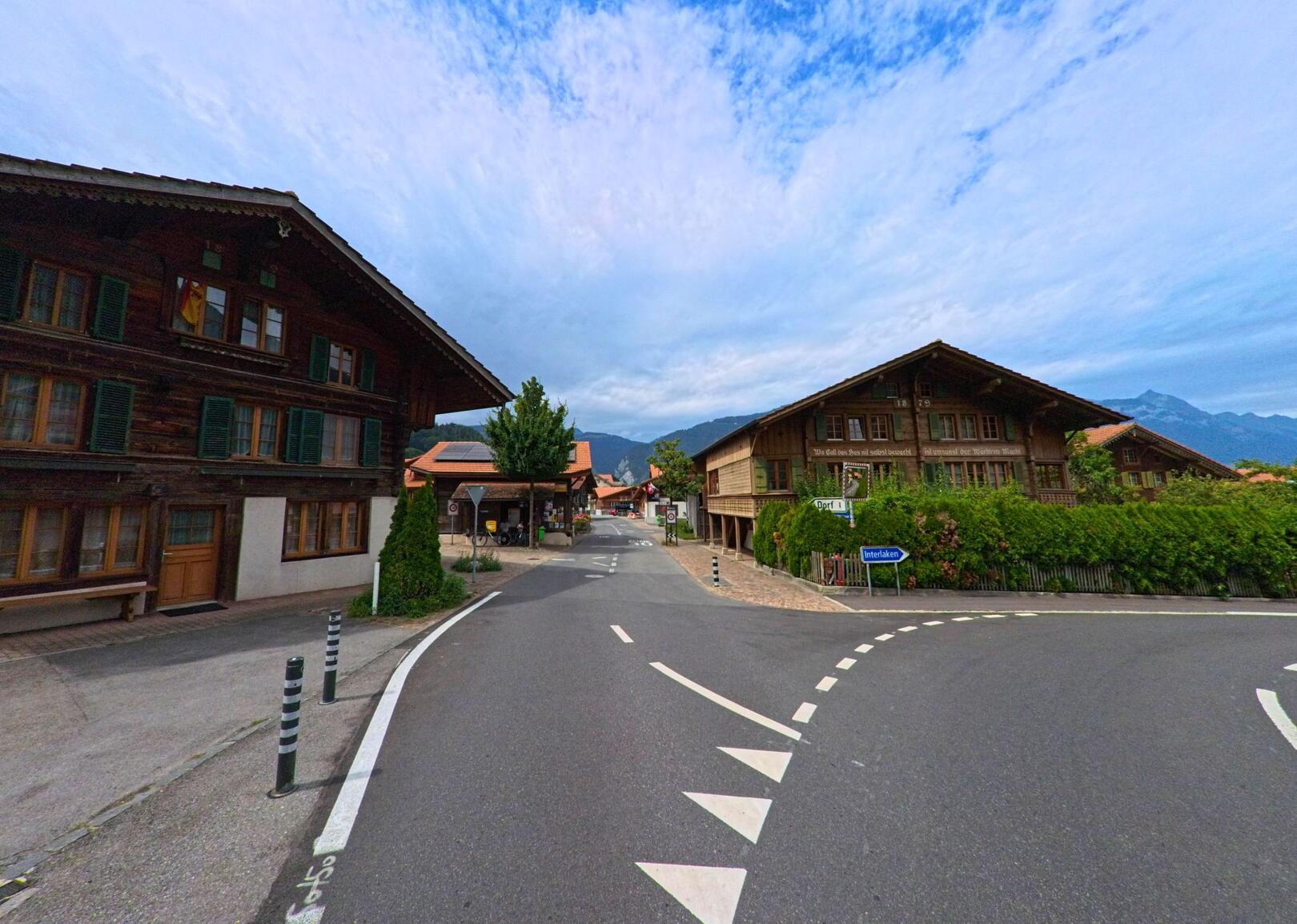 In Wilderswil (3812, Kanton Bern) mündet eine kleine Strasse zwischen traditionellen Holzhäusern in eine Kreuzung mit Blick auf die Berge im Hintergrund.