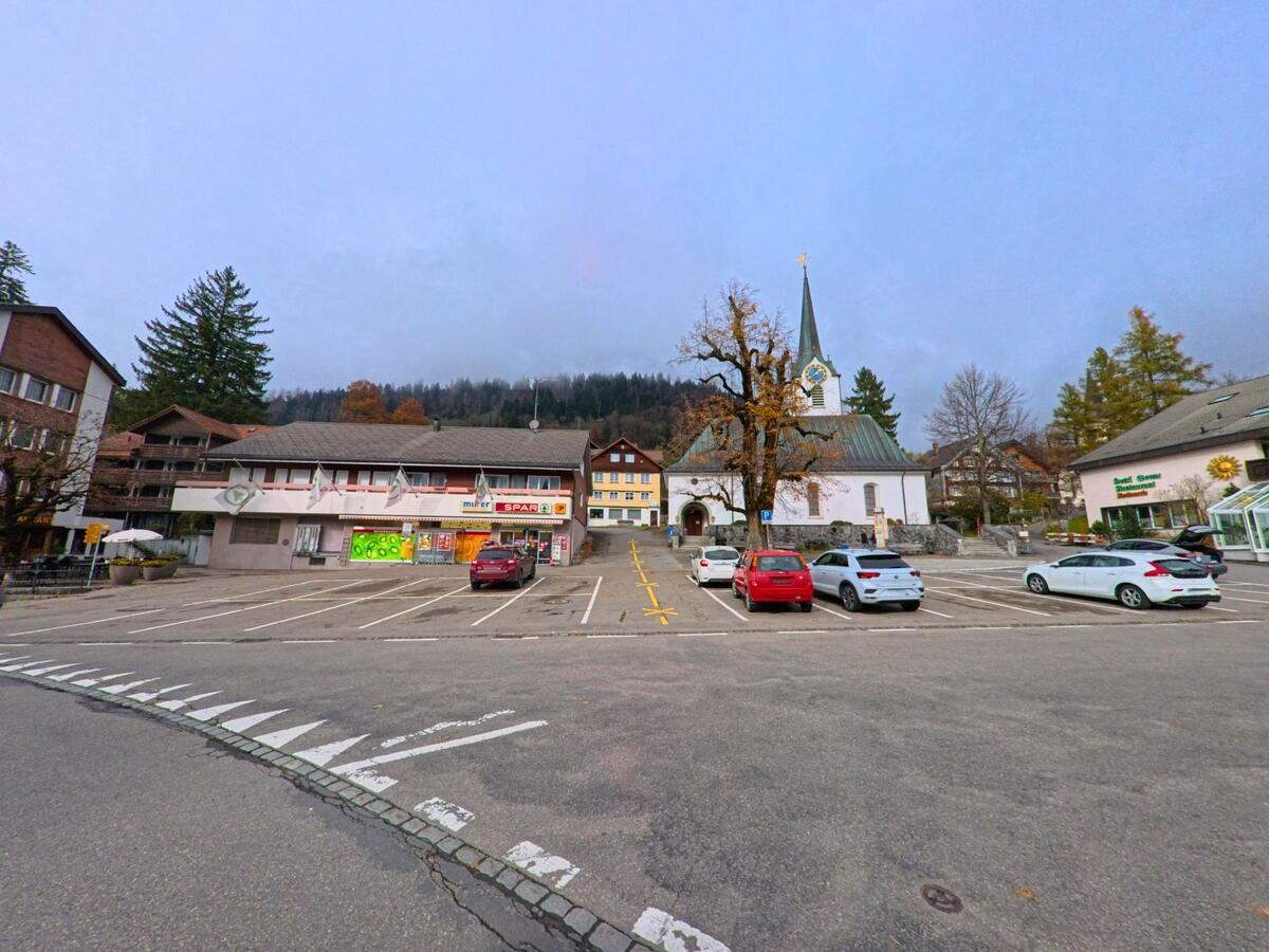 Vor Kirche und Geschäftszeile erstreckt sich ein zentraler Parkplatz im Ortskern von Wildhaus-Alt St. Johann (9656, Kanton St. Gallen) mit Blick auf bewaldete Hänge.