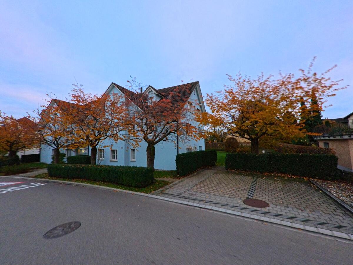 Vor einem Mehrfamilienhaus mit hellem Fassadenanstrich stehen herbstlich gefärbte Bäume entlang der Strasse in Wilen (9535, Kanton Thurgau) und prägen das ruhige Wohnumfeld.