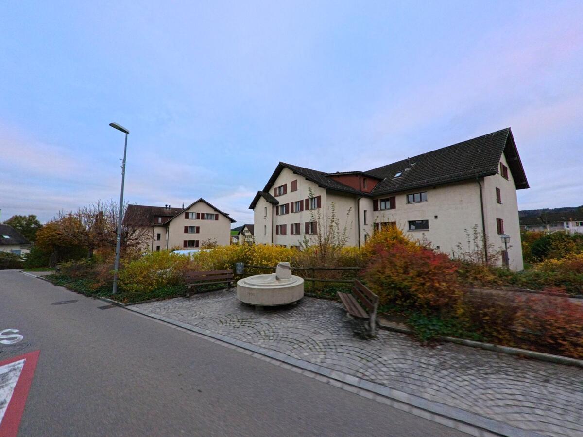 Ein kleiner Platz mit Sitzbänken und einem steinernen Brunnen liegt zwischen Wohnbauten und herbstlicher Bepflanzung in Wilen (9535, Kanton Thurgau) an einer ruhigen Quartierstrasse.