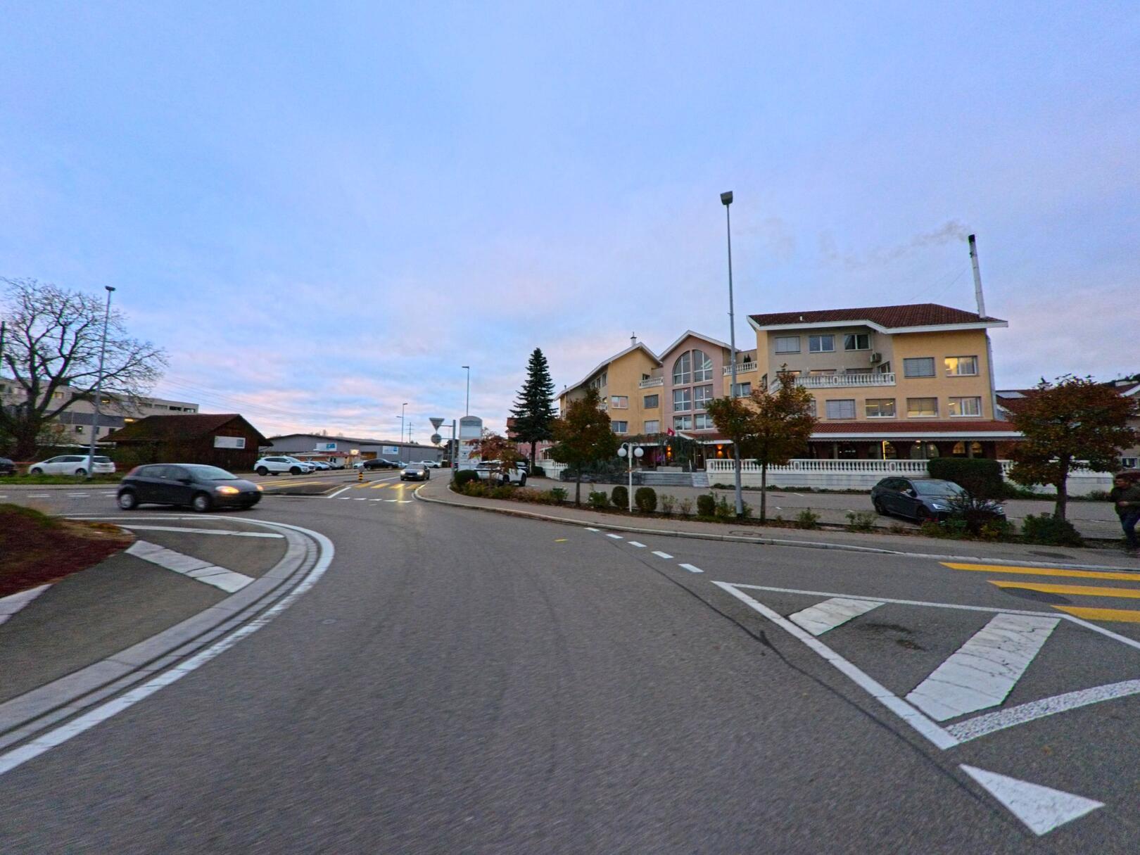 An einer verkehrsberuhigten Kreuzung mit Mittelinsel zeigt sich das Ortsbild von Wilen (9535, Kanton Thurgau) mit Wohnbauten und vorbeifahrenden Fahrzeugen in der Abenddaemmerung.