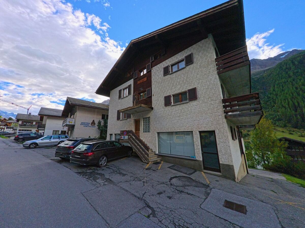 Ein Gebaeude mit Balkonen und braunen Fensterlaeden steht in Wiler (3918, Kanton Wallis) an einer leicht ansteigenden Strasse, vor dem mehrere Autos parken.