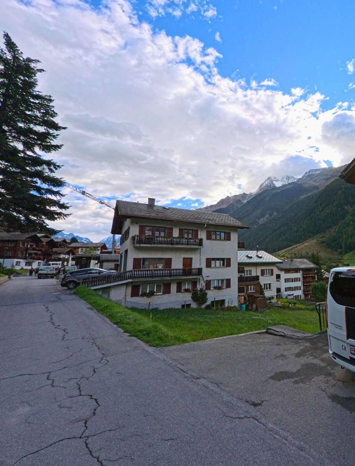 Ein mehrstoekiges Wohnhaus mit Balkonen und braunen Fensterlaeden steht in Wiler (3918, Kanton Wallis) am Strassenrand mit Blick auf die umliegenden Berge.