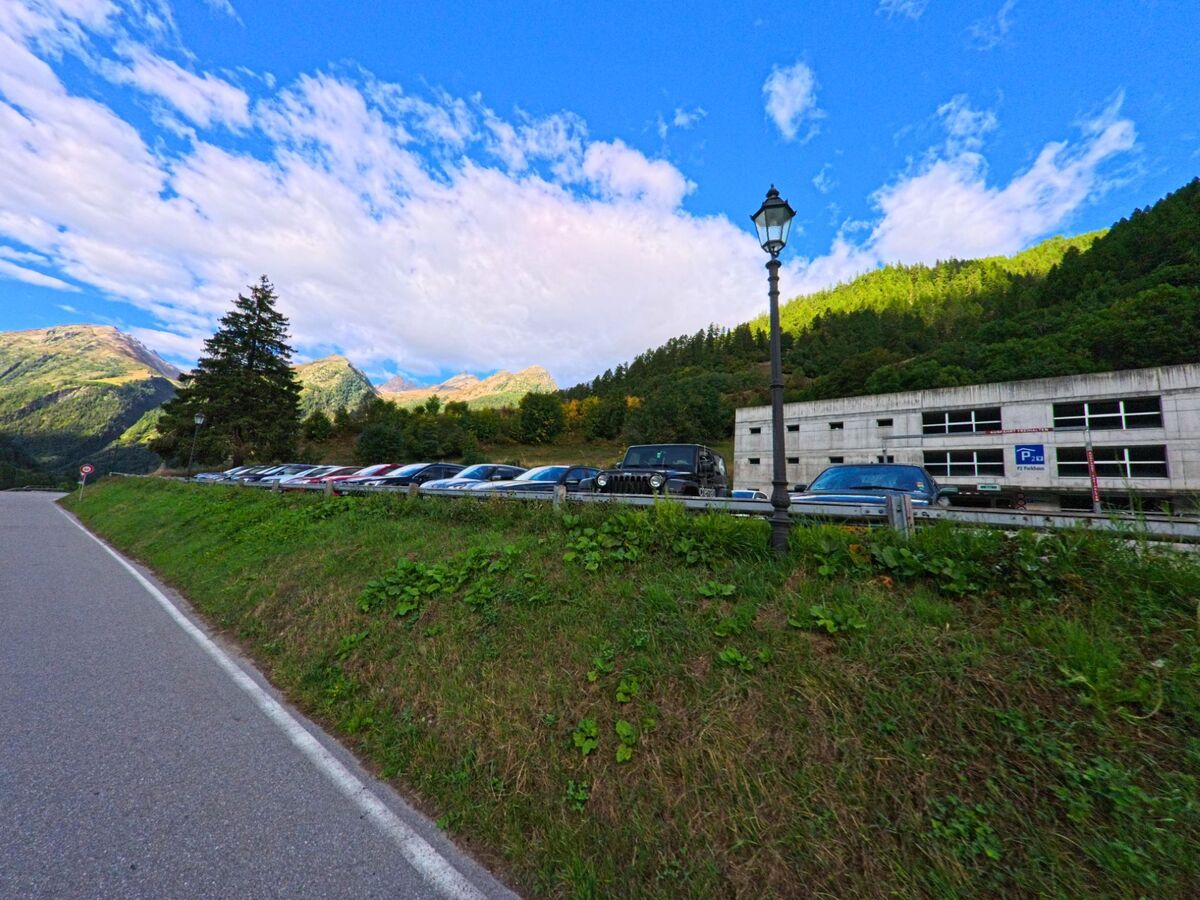 Ein Parkplatz am Ortsrand von Wiler (3918, Kanton Wallis) liegt neben einem modernen Gebaeude, dahinter erheben sich bewaldete Haenge und Berggipfel.