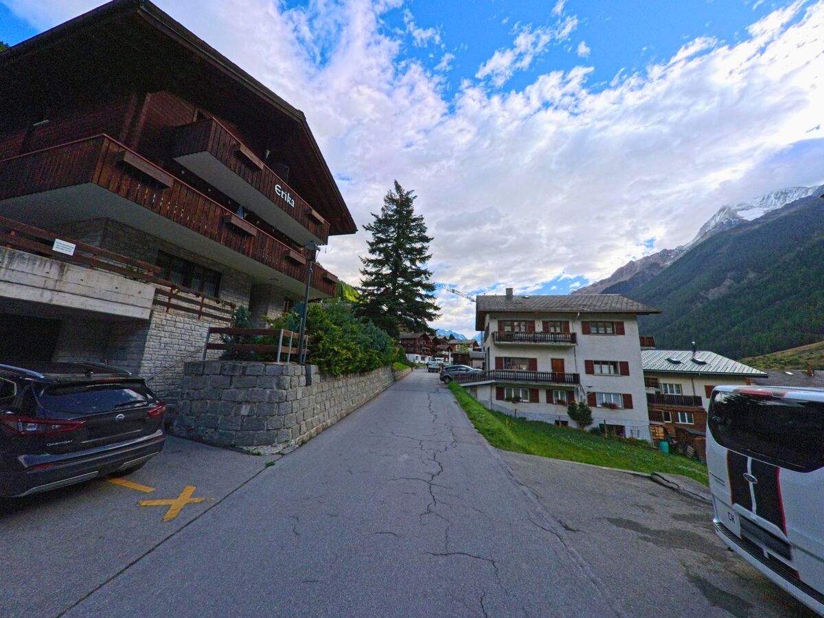 Eine schmale Strasse fuehrt in Wiler (3918, Kanton Wallis) zwischen Chalets und Mehrfamilienhaeusern bergauf, im Hintergrund sind verschneite Gipfel sichtbar.