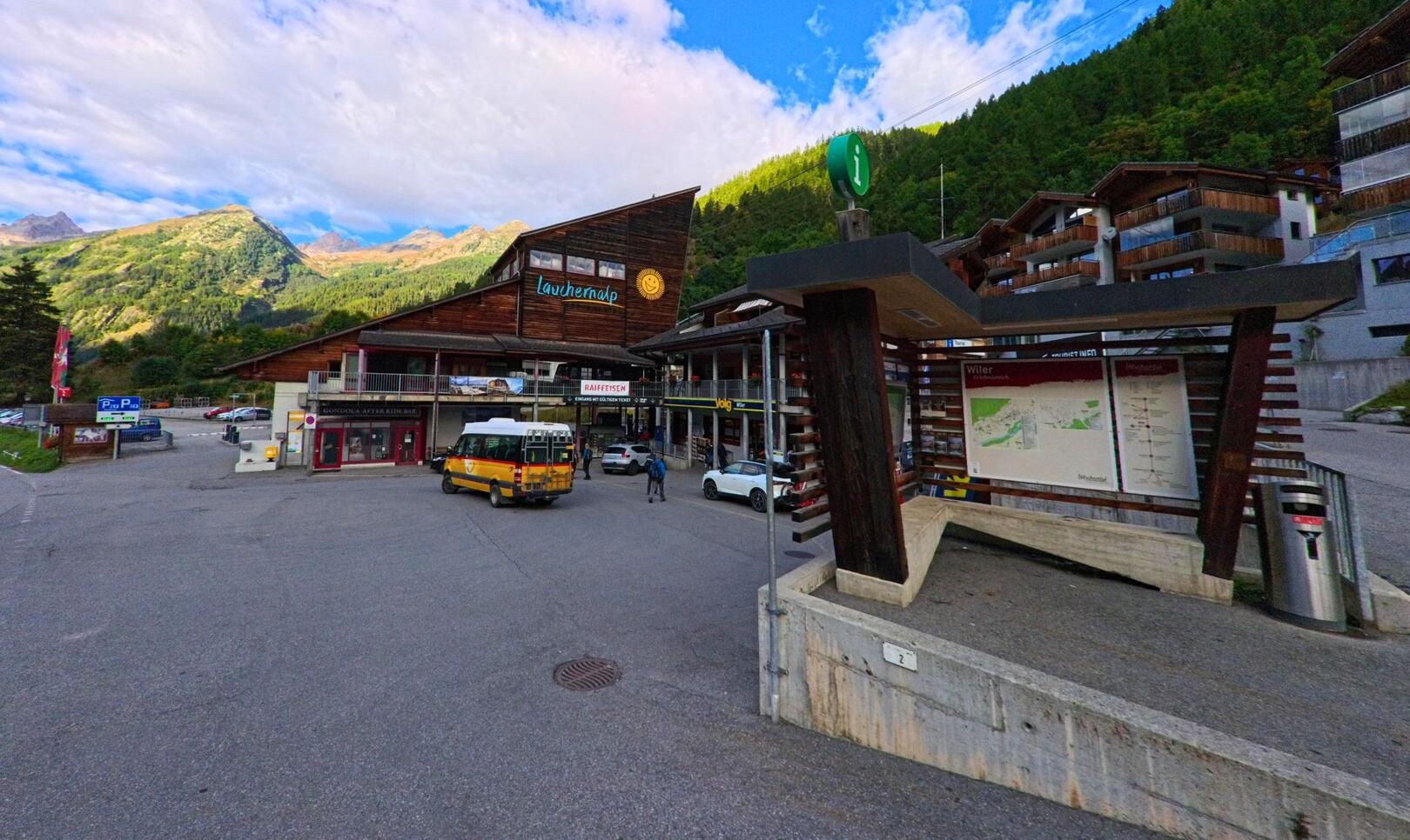 Am Eingang zur Talstation in Wiler (3918, Kanton Wallis) parken ein Postauto und mehrere Fahrzeuge, während dahinter die Berglandschaft des Lötschentals sichtbar ist.