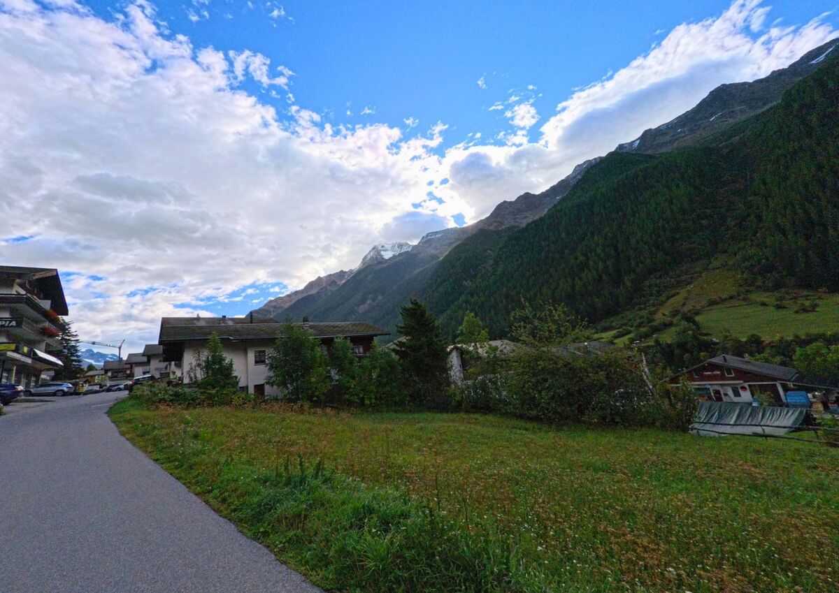 Am Rand von Wiler (3918, Kanton Wallis) fuehrt eine Strasse an Wiesen und Haeusern vorbei, im Hintergrund erheben sich bewaldete Haenge und schneebedeckte Gipfel.