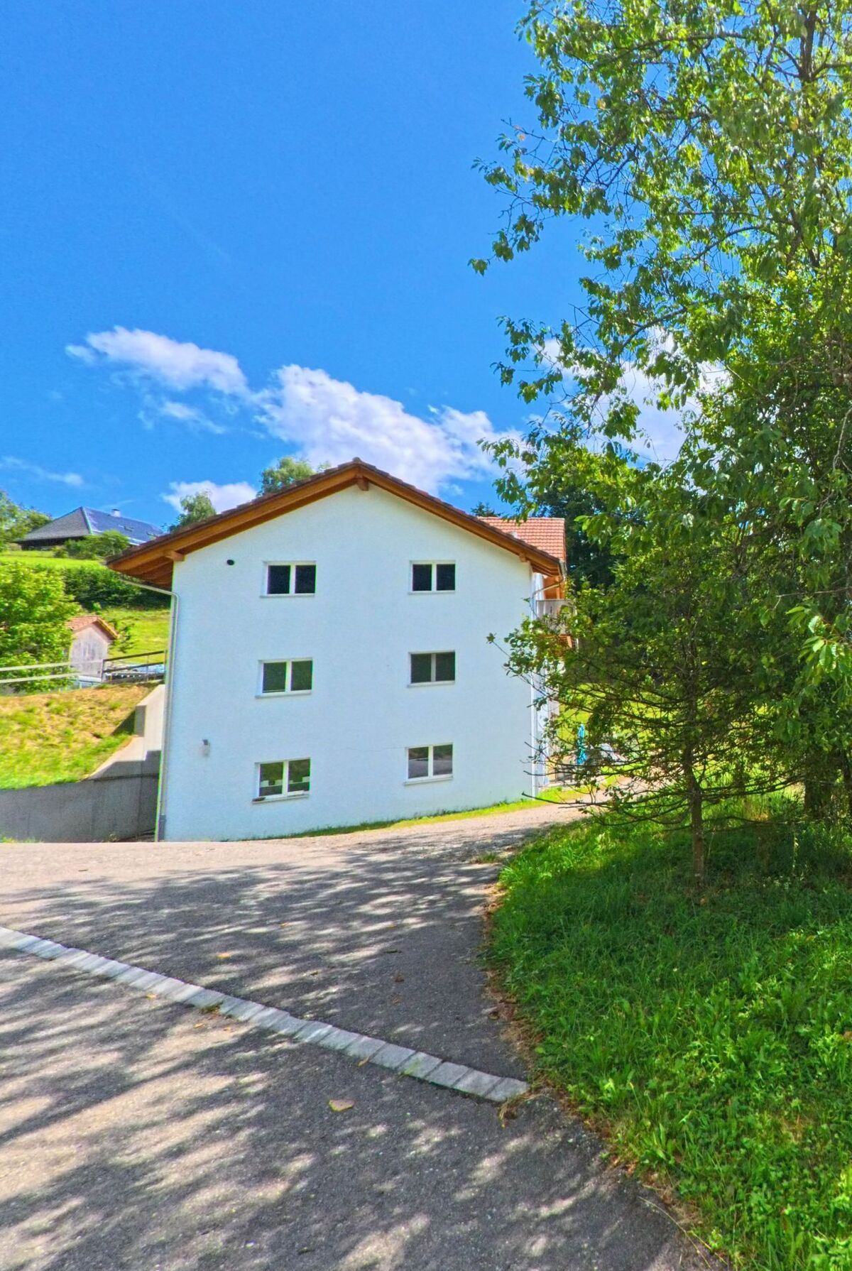 In Wiliberg (5058, Kanton Aargau) steht ein schlichtes, weisses Wohnhaus mit Satteldach an einem leicht abschuessigen Weg, umgeben von Wiesen und Baumen.