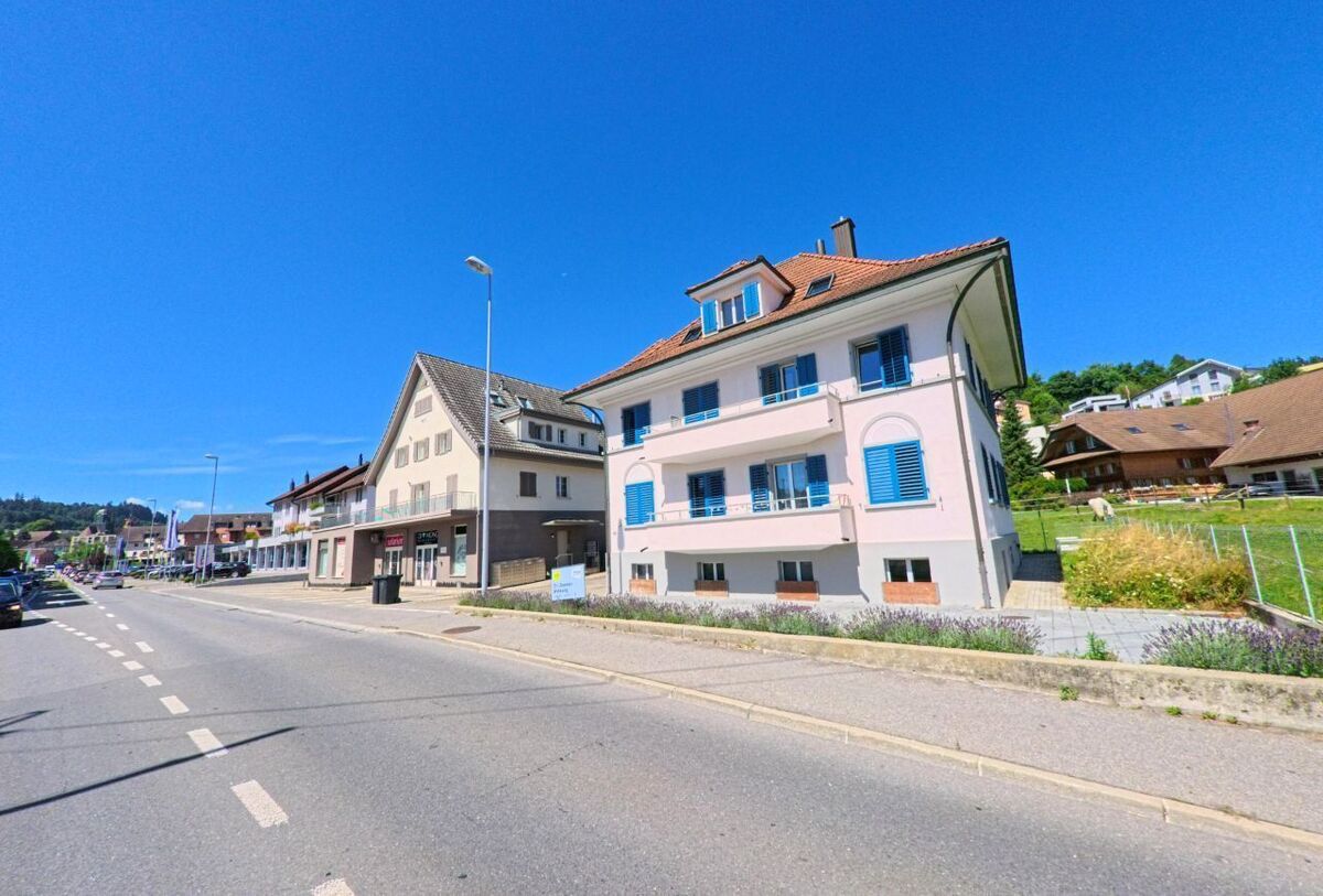 Entlang einer Hauptstrasse in Willisau (6130, Kanton Luzern) stehen Wohn- und Geschaeftshaeuser mit farbigen Fensterlaeden, im Hintergrund sind Einfamilienhaeuser am Hang zu sehen.