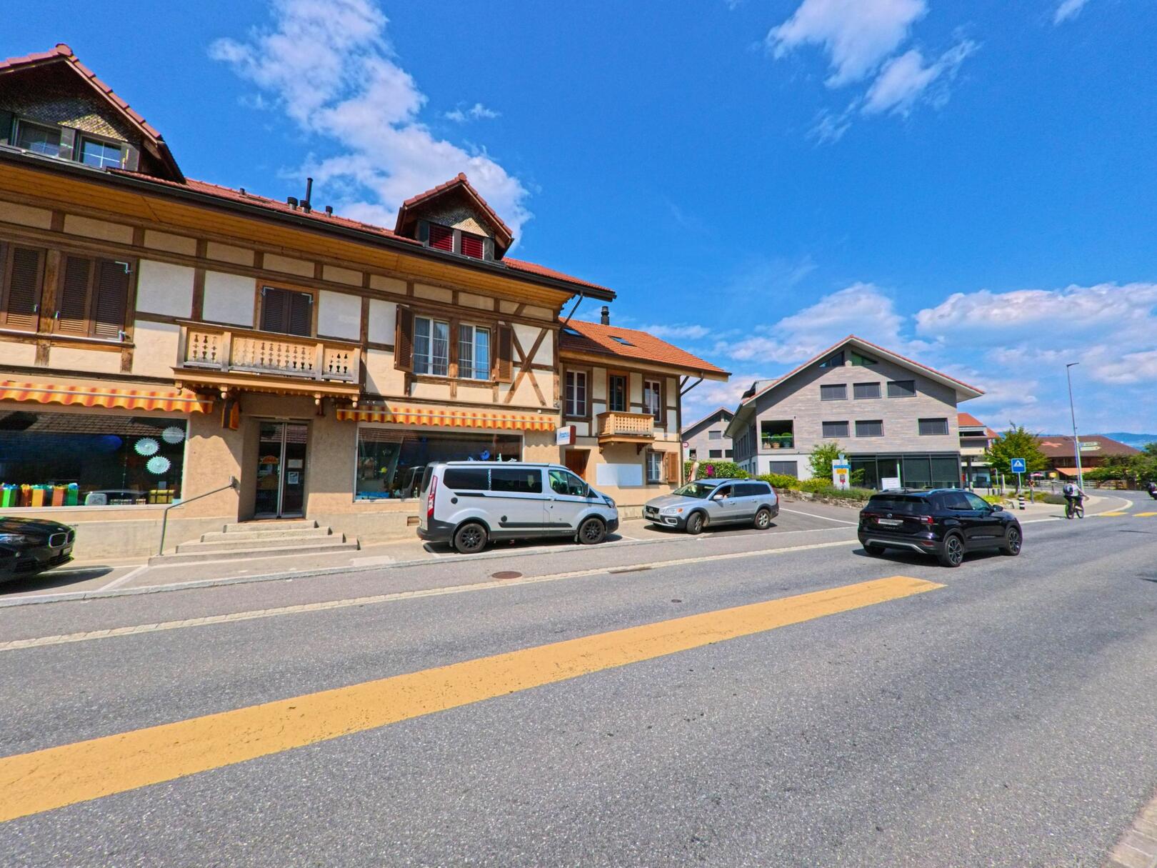 In der Gemeinde Wimmis (3752, Kanton Bern) stehen ein traditionelles Gebäude mit Holzfassade und ein moderner Neubau einander gegenüber, getrennt durch eine verkehrsreiche Strasse bei sonnigem Wetter.