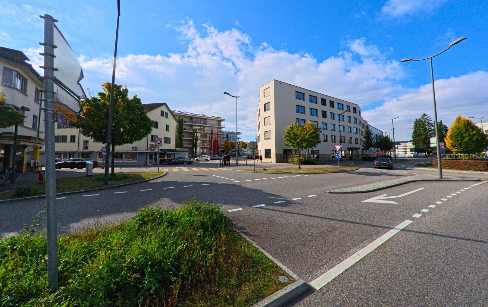 An einer belebten Kreuzung in Windisch (5210, Kanton Aargau) stehen moderne Wohnhäuser und ein kleiner Kreisel unter blauem Himmel.
