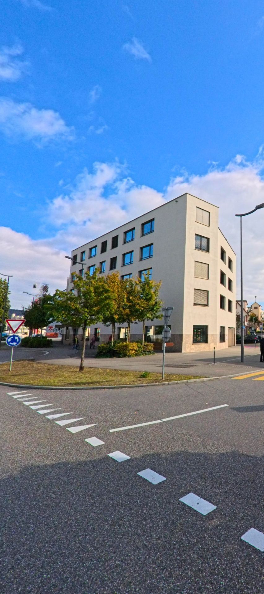 Ein modernes Eckgebaeude praegt in Windisch (5210, Kanton Aargau) den kleinen Kreisel, umgeben von jungen Baumen und klar markierten Fahrbahnen.