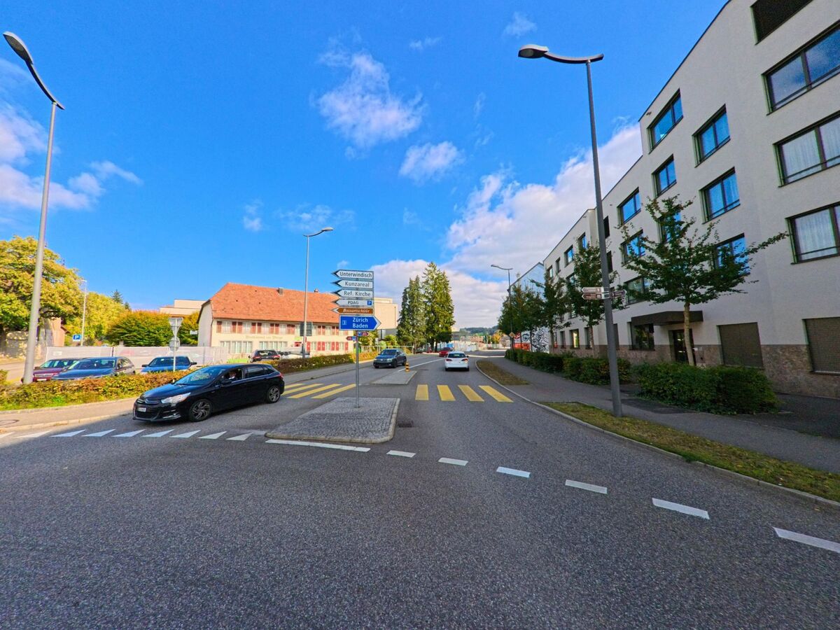 Ein breiter Strassenabschnitt in Windisch (5210, Kanton Aargau) fuehrt zwischen modernen Wohnhaeusern und Wegweisern vorbei, waehrend Autos den Zebrastreifen passieren.