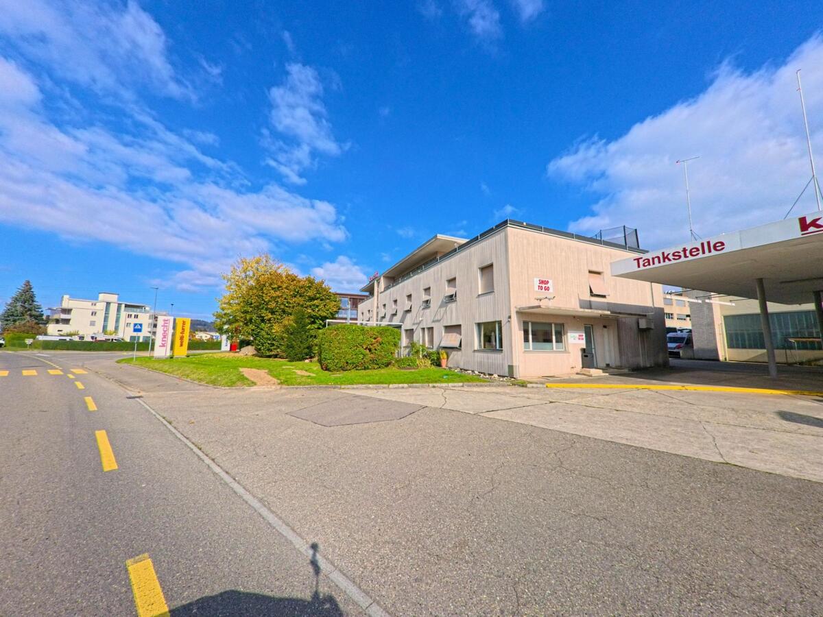 Neben einer Tankstelle verläuft in Windisch (5210, Kanton Aargau) eine breite Strasse, gesaeumt von schlichten Gewerbegebaeuden und gruener Bepflanzung.
