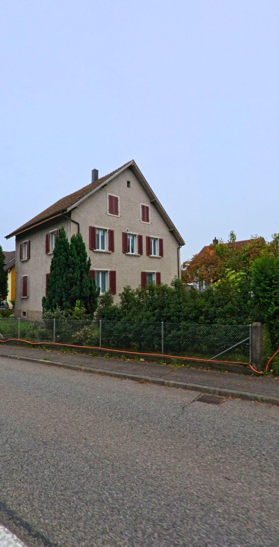 Ein schlichtes Wohnhaus mit roten Fensterlaeden steht in Windisch (5210, Kanton Aargau) an einer ruhigen Strasse, umgeben von dichter Begruenung.