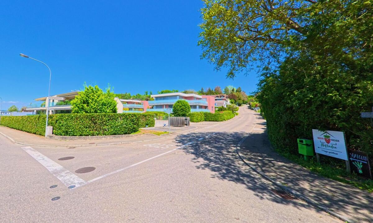 Am Gehrenweg in Winkel, PLZ 8185 im Kanton Zürich, führen gepflegte Quartierstrassen vorbei an modernen Wohnbauten und einem Hofladenhinweis unter schattigen Bäumen.