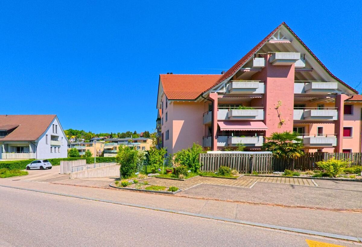 In Winkel, PLZ 8185 im Kanton Zürich, steht ein rosa Mehrfamilienhaus mit auffälligen Balkonen und Sonnenuhr an einer ruhigen Quartierstrasse.