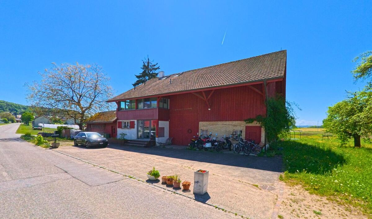 In Winkel, PLZ 8185 im Kanton Zürich, steht ein rotes Wohn- und Wirtschaftsgebäude mit angeschlossener Garage und Fahrrädern am Rand eines offenen Feldes.
