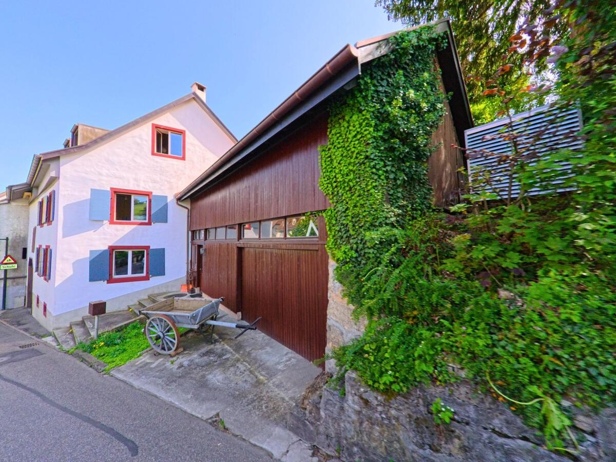 In einer Seitenstrasse von Wintersingen (4451, Kanton Baselland) steht ein Haus mit blauen Fensterlaeden neben einer von Efeu bewachsenen Holzscheune, vor der ein alter Handwagen abgestellt ist.