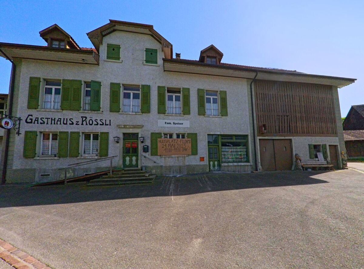 In Wintersingen (4451, Kanton Baselland) steht das Gasthaus zum Roessli mit gruuen Fensterlaeden und einem Veranstaltungsplakat an der Fassade auf einem gepflasterten Platz.