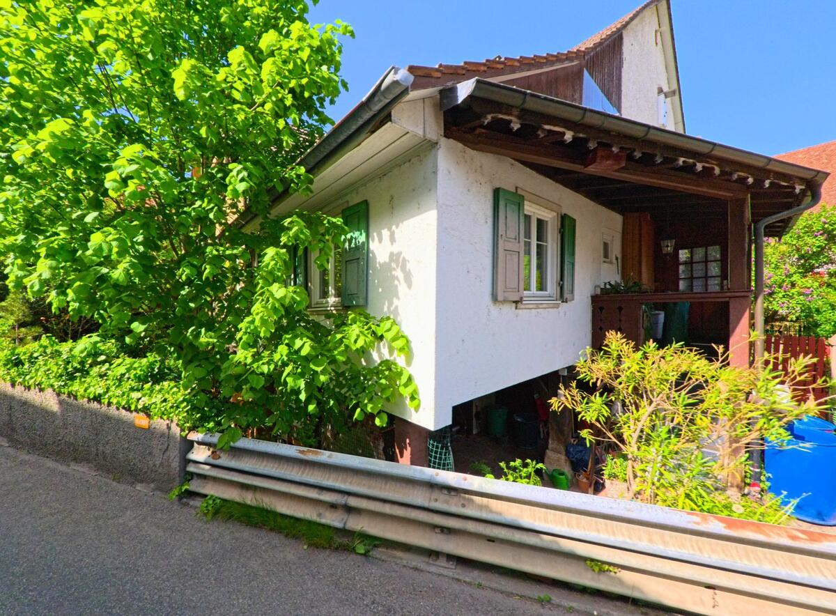 Ein kleines Wohnhaus mit gruenen Fensterlaeden und ueberdachter Veranda steht in Wintersingen (4451, Kanton Baselland) direkt an einer schmalen Strasse mit Leitplanke.