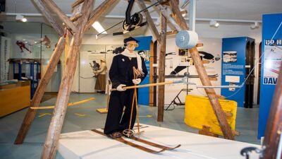 Im Wintersportmuseum in Davos Platz (7270, Kanton Graubünden) zeigt eine Ausstellung historische Skiausrüstungen und eine Figur in alter Skikleidung unter einer Holzseilbahnkonstruktion.