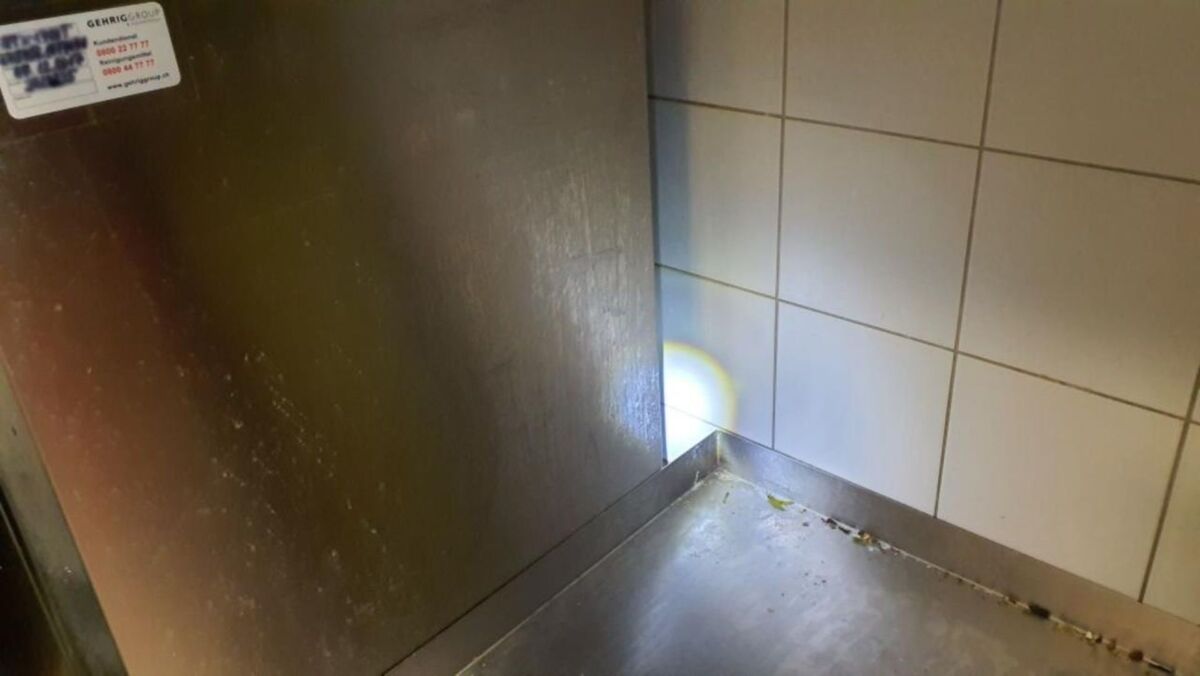  In einer Küche in Winterthur (8049, Kanton Zürich) beleuchtet eine Taschenlampe eine schmutzige Ecke zwischen Edelstahlgerät und gefliester Wand, in der sich Verunreinigungen gesammelt haben.