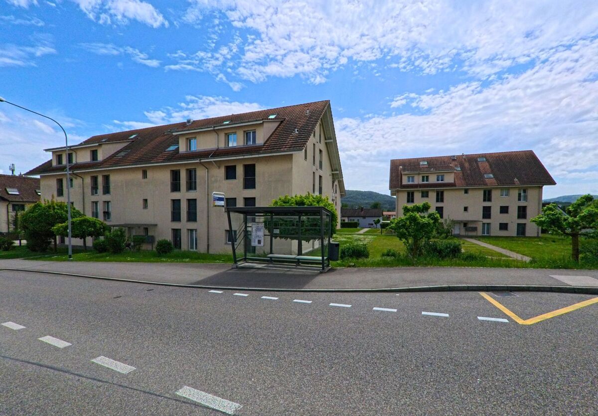 Vor mehreren hellen Mehrfamilienhäusern in Winznau (4652, Kanton Solothurn) steht eine überdachte Bushaltestelle an einer ruhig gelegenen Strasse mit gepflegtem Grünbereich.