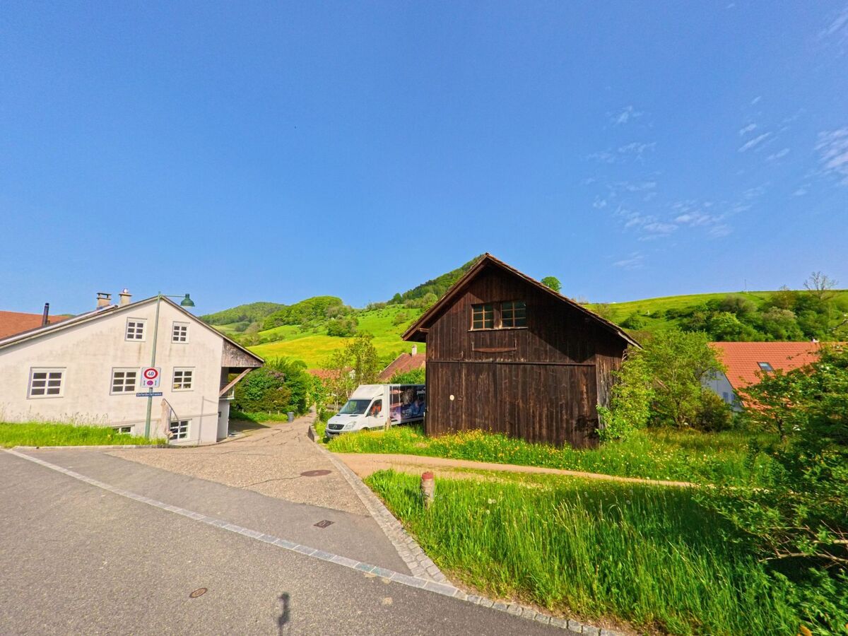 In Wisen (4634, Kanton Solothurn) treffen sich eine schmale Gasse und eine Hauptstrasse neben einem alten Holzschuppen mit Blick auf die grünen Hügel.
