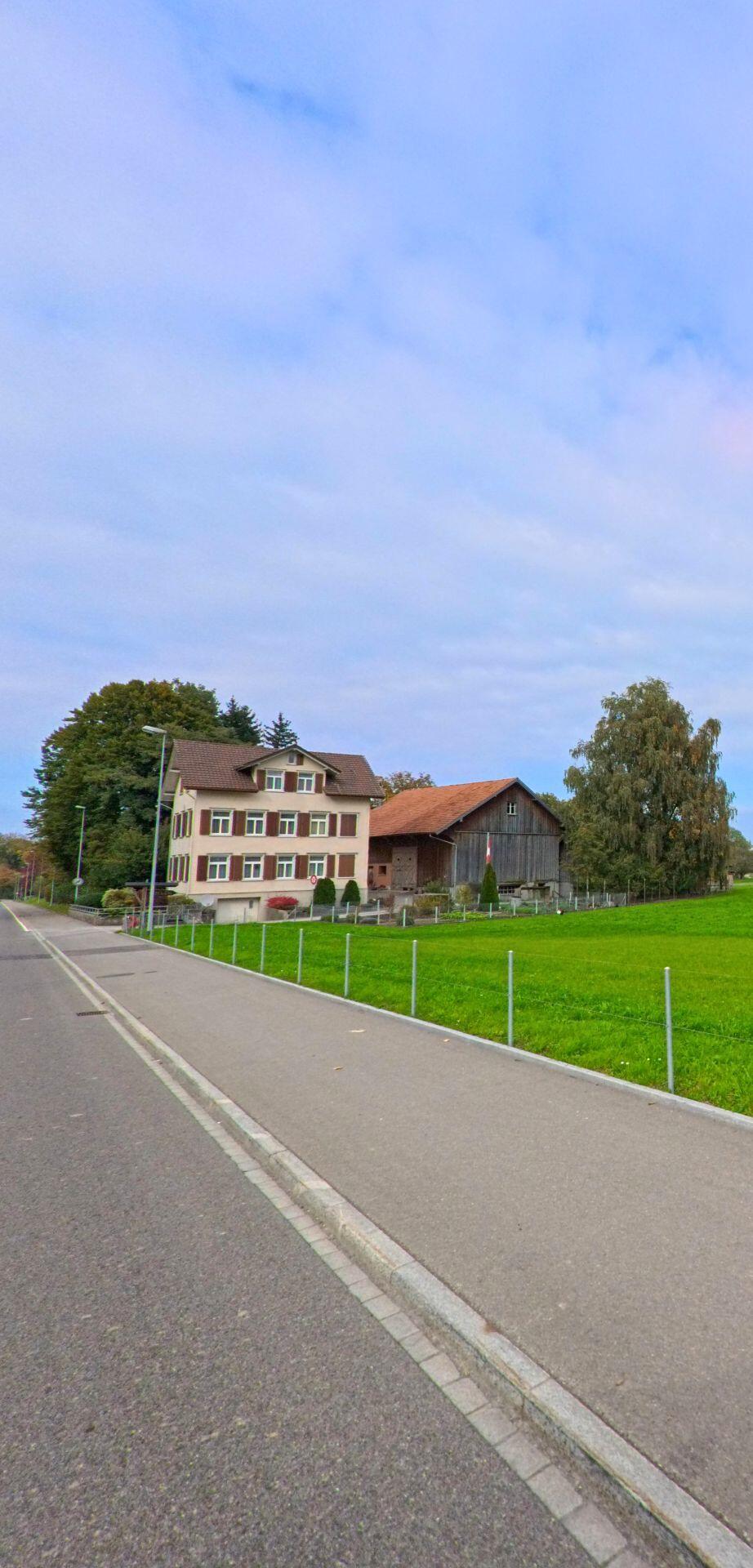 Entlang eines Gehwegs in Wittenbach (9300, Kanton St. Gallen) stehen ein Wohnhaus und eine Scheune neben gepflegten Gruenflaechen und Baumbestand.