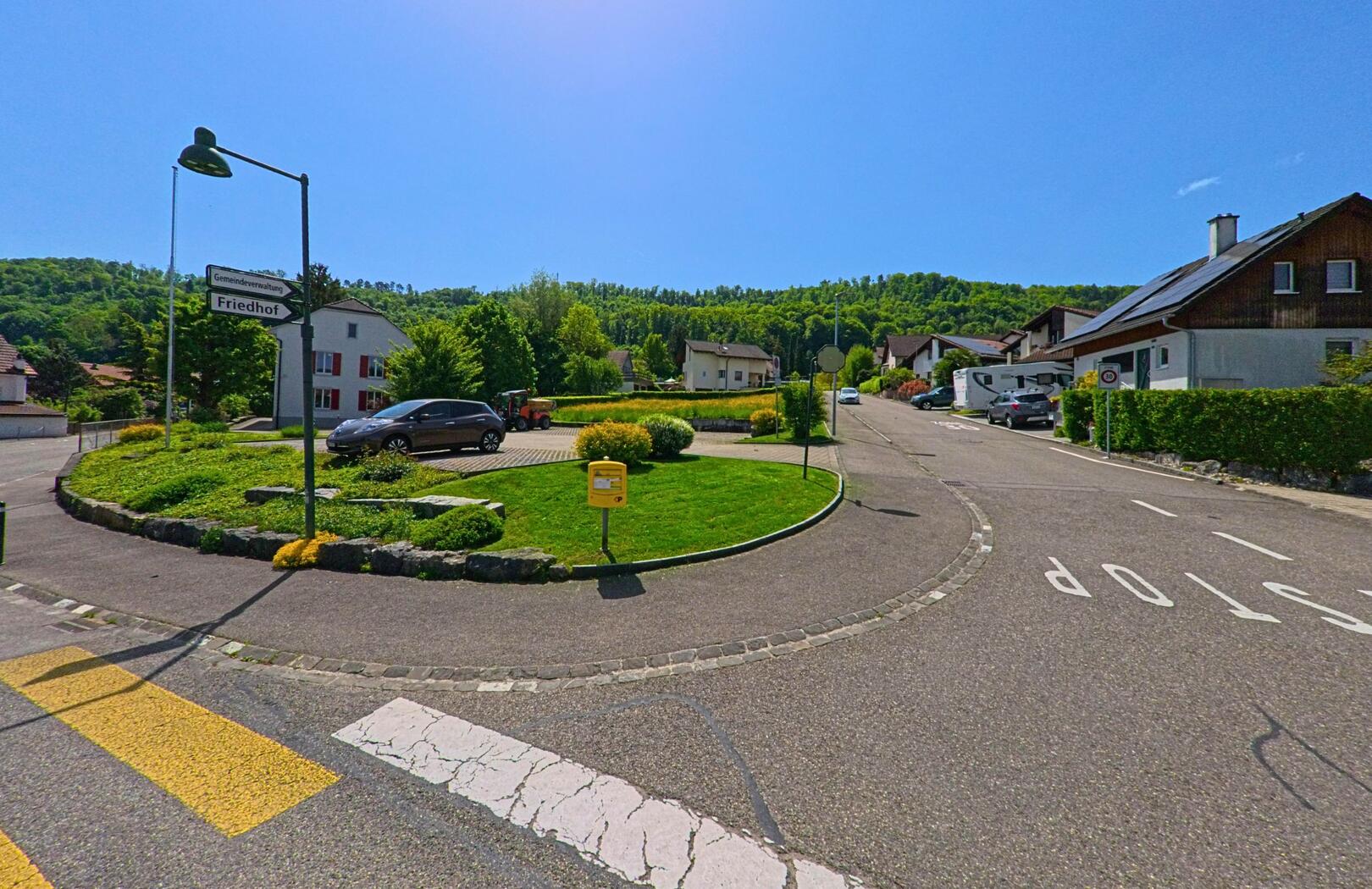 In Witterswil (4108, Kanton Solothurn) markiert ein kleiner bepflanzter Kreisverkehr mit Wegweisern zur Gemeindeverwaltung und zum Friedhof den Beginn einer ruhigen Wohnstrasse.