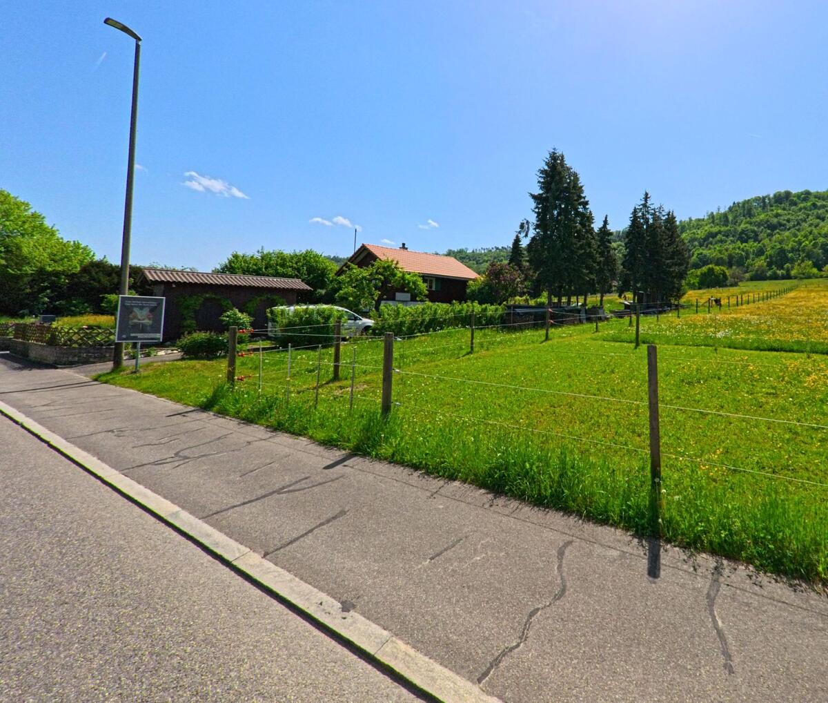 In Witterswil (4108, Kanton Solothurn) verläuft eine Strasse entlang eingezäunter Weiden und einzelner Bauernhäuser am Rand eines bewaldeten Hügels.