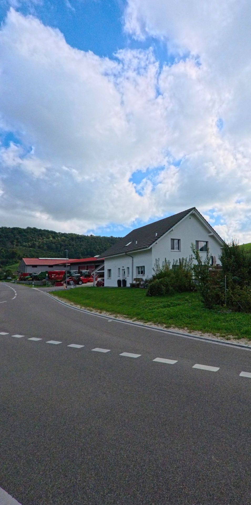 In Wittnau (5064, Kanton Aargau) steht ein weisses Einfamilienhaus am Rand einer kurvigen Strasse, neben der ein landwirtschaftlicher Betrieb mit roten Daechern liegt.