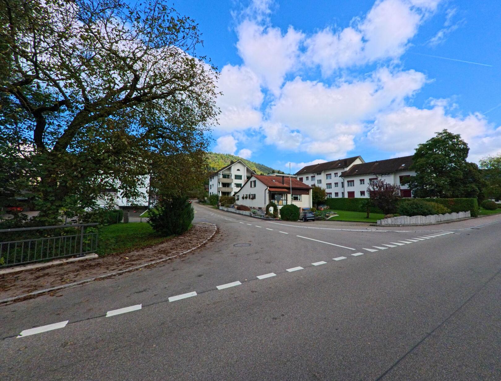 In Wittnau (5064, Kanton Aargau) fuehrt eine breite Strasse an gepflegten Wohnhaeusern vorbei, waehrend im Hintergrund sanfte Huegel unter blauem Himmel sichtbar sind.
