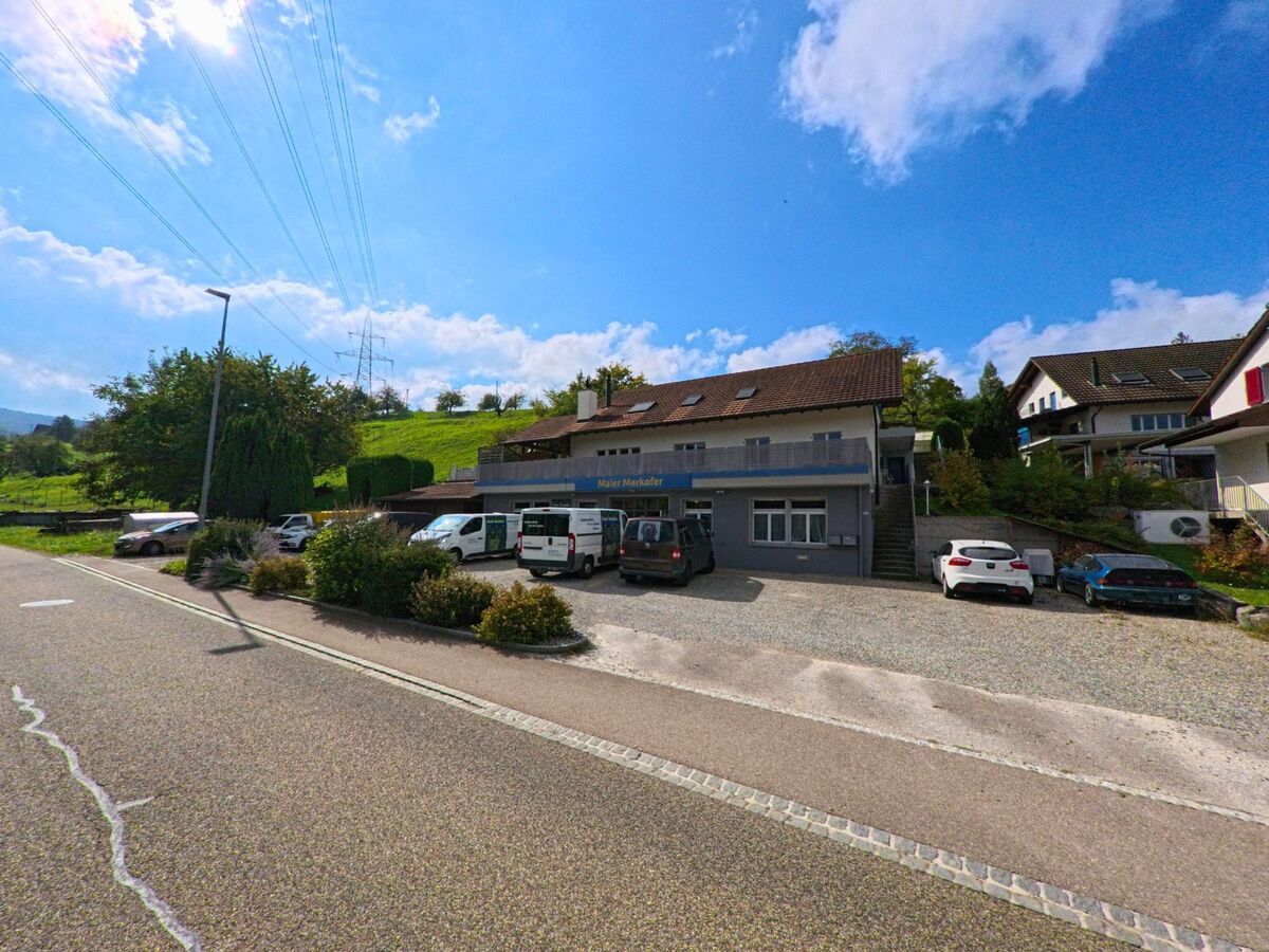 Am Hangrand von Woelflinswil (5063, Kanton Aargau) stehen mehrere Fahrzeuge vor einem Gewerbebau, waehrend sich dahinter gruene Wiesen und Stromleitungen erstrecken.