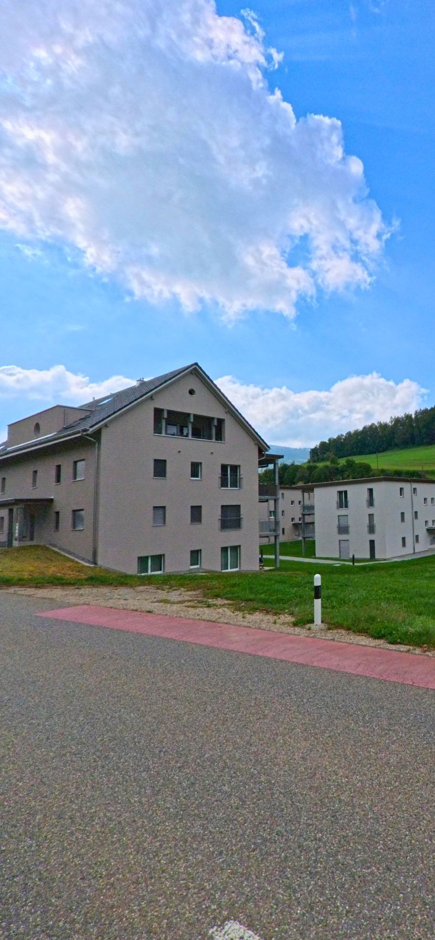 Mehrgeschossige Neubauten stehen in Woelflinswil (5063, Kanton Aargau) am sanft ansteigenden Ortsrand vor einer gruener Huegellandschaft unter lichtem Himmel.