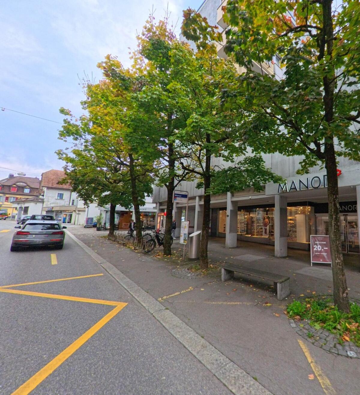 In der Einkaufsstrasse von Wohlen (5610, Kanton Aargau) stehen Bäume vor dem Manor-Gebäude, daneben parken Autos und Fahrräder am Strassenrand.