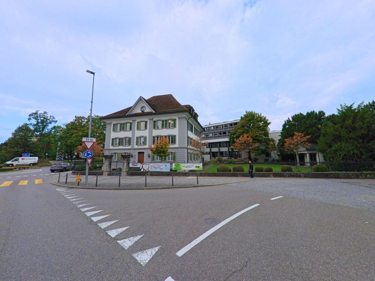 Ein klassisches Gebäude mit grünen Fensterläden steht an einem Kreisel in Wohlen (5610, Kanton Aargau), umgeben von gepflegten Bäumen und Rasenflächen.