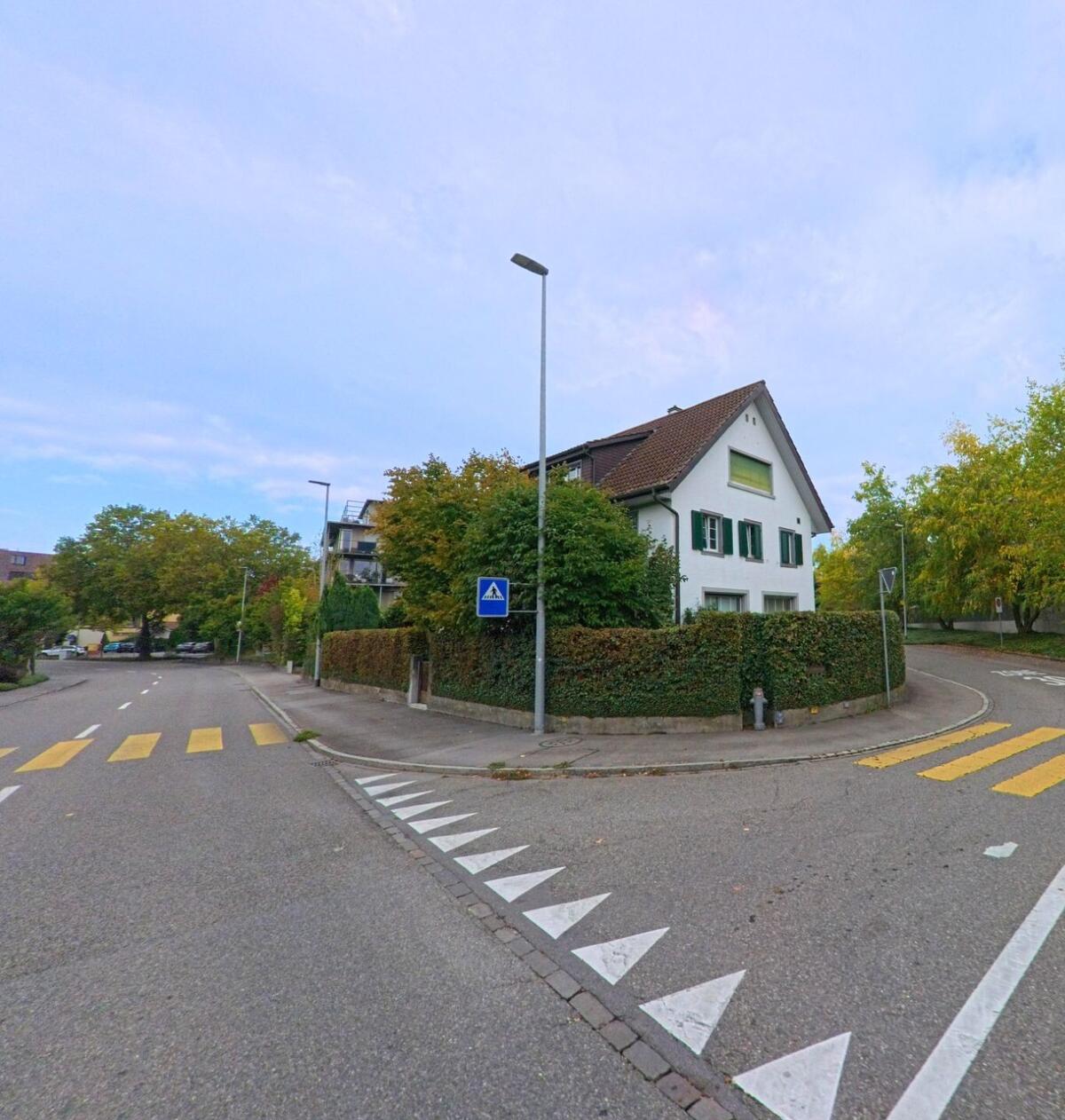An einer Kreuzung in Wohlen (5610, Kanton Aargau) steht ein weisses Wohnhaus mit grünen Fensterläden, umgeben von Bäumen und Strassen mit Fussgängerstreifen.