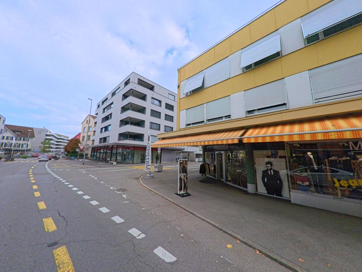 In Wohlen (5610, Kanton Aargau) verläuft eine Strasse mit modernen Wohnhäusern und einem Bekleidungsgeschäft im Erdgeschoss unter orangefarbenen Markisen.